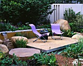 RSPB GARDEN  CHELSEA 2002. WATER FEATURE: RILL WITH WATERFALL SURROUNDED BY BOULDERS  TERRACE WITH LILAC CHAIRS