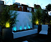 ROOF GARDEN LIT UP AT NIGHT WITH DECKING  WHITE WATER FEATURE  CHAIRS AND GALVANISED POTS PLANTED WITH BAMBOOS. DESIGNERS: PATRICK WYNNIAT - HUSEY AND PATRICK CLARKE