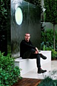 GARDEN DESIGNER STEPHEN WOODHAMS SITS ON THE WATER FEATURE. SANCTUARY GARDEN SPONSORED BY MERRILL LYNCH AT THE CHELSEA FLOWER SHOW 2002: DESIGNED BY STEPHEN WOODHAMS: