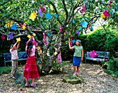 DESIGNER CLARE MATTHEWS: CHILDRENS PARTY -  GIRLS WITH SELF MADE PARTY HATS HANGING FLOWER MOBILES ONTO AN APPLE TREE IN THE GRAVEL GARDEN