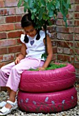 DESIGNER: CLARE MATTHEWS - FRAGRANT TREE SEAT - GIRL SITTING ON PINK PAINTED CAR TYRES PLANTED WITH A FIG TREE AND THYMES