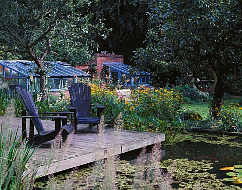 VIEW_OVER_LILY_POOL_TO_WOODEN_DECK_WITH_ADIRONDACK_CHAIRS_IN_THE_BACKGROUND_ARE_TWO_GREENHOUSES_GREY