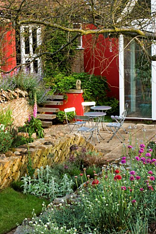 VIEW_TO_THE_PATIO_WITH_METAL_TABLE_AND_CHAIRS__RED_WALLS_AND_ANGLED_DRYSTONE_WALLING_WINGWELL_NURSER