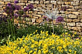 A DELICATE GLASS SCULPTURE BY NEIL WILKINS IN A BORDER BESIDE A STONE WALL WITH ALLIUMS AND HELIANTHEMUM. WINGWELL NURSERY   RUTLAND