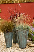 GROUP OF THREE CONTAINERS PLANTED WITH ORNAMENTAL GRASSES CAREX  PENNISETUM AND FESTUCA GLAUCA STAND IN GRAVEL IN FRONT OF RED WALL. CONTAINERS BY GREEN INTERIORS.