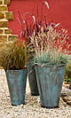 GROUP OF THREE CONTAINERS PLANTED WITH ORNAMENTAL GRASSES CAREX  PENNISETUM AND FESTUCA GLAUCA STAND IN GRAVEL IN FRONT OF RED WALL. CONTAINERS BY GREEN INTERIORS.