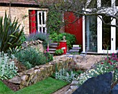 WINGWELL NURSERY  RUTLAND: VIEW TO THE BACK OF THE HOUSE WITH RED WALL  STACHYS  PHORMIUM   METAL TABLE AND CHAIRS. PATIO