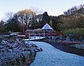 GARDEN DESIGNED BY DUNCAN HEATHER: THE SUMMERHOUSE AND WOODEN BRIDGE ACROSS THE POOL IN WINTER WITH CORNUS AND GUNNERA MANICATA