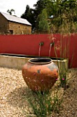 RICKYARD BARN  NORTHAMPTONSHIRE: THE GRAVEL COURTYARD IN JUNE WITH LARGE TERRACOTTA CONTAINER  RED WALL  WATER SPOUTS AND TROUGH