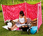 DESIGNER CLARE MATTHEWS: WIND BREAK SCREEN PROJECT - CLARE SITS IN THE WIND BREAK IN LAWN WITH CUSHIONS  BARBEQUE AND TRAY WITH DRINKS