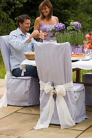 DESIGNER_CLARE_MATTHEWS_CLARE_AND_MAN_BY_TABLE_WITH_CHAIR_COVERED_WITH_PALE_BLUE_SHEET_WITH_WHITE_RI