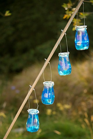 DESIGNER_CLARE_MATTHEWS__LANTERN_STAND_PROJECT_WOODEN_ROD_WITH_BLUE_GLASS_CANDLE_JARS_HANGING_OFF