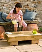 DESIGNER: CLARE MATTHEWS - LOW SLEEPER TABLE PROJECT: GIRL SITTING ON WOODEN BENCH POURING ORANGE JUICE FROM LOW SLEEPER TABLE ON PATIO