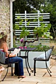 DESIGNER: CLARE MATTHEWS - MOBILE FRUIT SCREEN - GIRL SITS READING AT TABLE WITH CONTAINER AND TRELLIS SCREEN WITH VINE AND STRAWBERRIES ELSANTA BESIDE A WALL ON PATIO