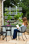DESIGNER: CLARE MATTHEWS - MOBILE FRUIT SCREEN - GIRL SITS READING AT TABLE WITH CONTAINER AND TRELLIS SCREEN WITH VINE AND STRAWBERRIES ELSANTA BESIDE A WALL ON PATIO