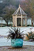 WOODPECKERS  WARWICKSHIRE  WINTER: COPPER CONTAINER WITH PHORMIUM ON BRICK CIRCLE WITH WOODEN SUMMERHOUSE BEHIND  IN FROST