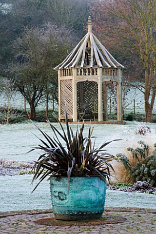 WOODPECKERS__WARWICKSHIRE__WINTER_COPPER_CONTAINER_WITH_PHORMIUM_ON_BRICK_CIRCLE_WITH_WOODEN_SUMMERH