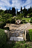PROVENCE  FRANCE. GARDEN OF MARCO NUCERA. GRAVEL GARDEN WITH WOODEN SCULPTURE  STONE WATER TROUGH  WOODEN PATH AND TREES CLIPPED BY MARCO NUCERA