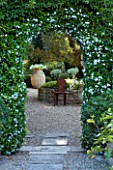DESIGNER MICHEL SEMINI  PROVENCE  FRANCE. VIEW THROUGH DOORWAY WITH TRACHELOSPERMUM JASMINOIDES TO GRAVEL COURTYARD WITH TERRACOTTA CONTAINER AND ORNATE METAL SEATS
