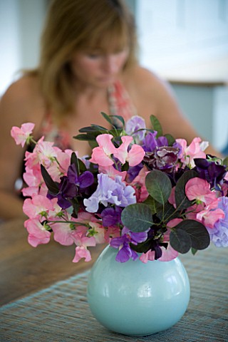 CLARE_MATTHEWS_HOUSE__DEVON_THE_KITCHEN_TABLE_WITH_BLUE_VASE_OF_SWEETPEAS__CLARE_IN_BACKGROUND