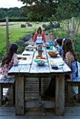 DESIGNER CLARE MATTHEWS: DEVON GARDEN. OUTDOOR SEATING AREA. WOODEN TABLE AND BENCHES ON PATIO. CLARE AND CHILDREN ABOUT TO EAT LUNCH