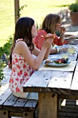 DESIGNER CLARE MATTHEWS: DEVON GARDEN. OUTDOOR SEATING AREA. WOODEN TABLE AND BENCHES ON PATIO. CLARE AND CHILDREN EATING LUNCH