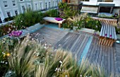 ROOF GARDEN HOLLAND PARK LONDON.DESIGNER CHARLOTTE ROWE.DECKED TERRACE WOODEN BENCHES PINK CUSHIONS BLUE GLASS GRAVEL  GAURA LINDHEIMERI WHIRLING BUTTERFLIES  STIPA TENUISSIMA