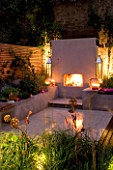 DESIGNER CHARLOTTE ROWE  LONDON: CHARLOTTE ROWES OWN GARDEN AT NIGHT - PORTUGUESE HONED LIMESTONE FLOORING  RENDERED RAISED BEDS AND FIREPLACE  PHILIPPE STARCK CHAIRS  TRELLIS