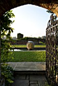 DAVID HARBER SUNDIALS: VIEW THROUGH A GATE AND ARCHWAY TO ETHER WATER FEATURE IN A POOL IN EARLY MORNING DAWN LIGHT WITH CHAMOMILE LAWN