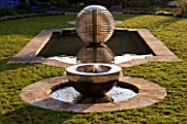 DAVID HARBER SUNDIALS: CHALICE SUNDIAL WATER FEATURE MADE OF MIRROR POLISHED STAINLESS STEEL SITS IN A POOL SURROUNDED BY CAMOMILE LAWN WITH ETHER WATER FEATURE BEHIND