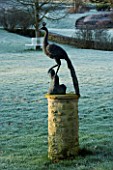 CERNEY HOUSE GARDEN  GLOUCESTERSHIRE:PEACOCK SCULPTURE ON A PEDESTAL ON THE LAWN IN FONT OF THE HOUSE IN FROST