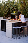 CONTEMPORARY FORMAL ROOF TERRACE/ GARDEN DESIGNED BY DATA NATURE ASSOCIATES: GIRL SITTING AT A TABLE BESIDE THE BARBEQUE