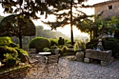PROVENCE  FRANCE: GARDEN OF NICOLE DE VESIAN  LA LOUVE: GRAVEL TERRACE BESIDE THE HOUSE AT DAWN WITH METAL TABLE AND CHAIRS  CLIPPED TOPIARY SHAPES AND AN OLD WELL