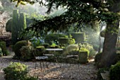 PROVENCE  FRANCE: GARDEN OF NICOLE DE VESIAN  LA LOUVE: GRAVEL TERRACE AT DAWN WITH METAL TABLE AND CHAIRS AND CLIPPED TOPIAREY SHAPES