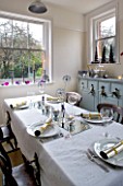 CLARE MATTHEWS CHRISTMAS HOUSE INTERIOR: THE KITCHEN WITH TABLE LAID FOR CHRISTMAS - PLATES WITH CRACKERS  CANDLES ON MIRROR AND IN WINDOWSILL  WINE GLASSES  BAUBLES  DRIED ALLIUMS