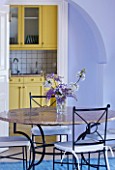 DESIGNER GINA PRICE - CORFU - VILLA ONEIRO - VIEW TO DINING AREA WITH TABLE AND JUG WITH WISTERIA AND BLOSSOM TO LEMON YELLOW KITCHEN BEYOND