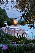CORFU  GREECE: DESIGNER: DOMINIC SKINNER - MEDITTERANEAN STYLE GARDEN  - VIEW TO SWIMMING POOL AT NIGHT WITH SUN LOUNGERS AND TERRACOTTA CONTAINER  LIGHTING