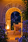 CORFU  GREECE: DESIGNER: DOMINIC SKINNER - MEDITTERANEAN STYLE GARDEN  - VIEW THROUGH TWO STONE ARCHES LIT UP AT NIGHT WITH TABLE  CHAIRS AND AGAPANTHUS