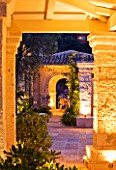 CORFU  GREECE: DESIGNER: DOMINIC SKINNER - MEDITTERANEAN STYLE GARDEN  - VIEW THROUGH ARCHES LIT UP AT NIGHT WITH PATHS  TABLE  CHAIRS AND AGAPANTHUS