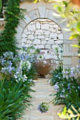 CORFU  GREECE: DESIGNER: DOMINIC SKINNER - MEDITTERANEAN STYLE GARDEN  - VIEW ALONG PATH THROUGH STONE ARCH TO FOCAL POINT TERRACOTTA CONTAINER AND AGAPANTHUS