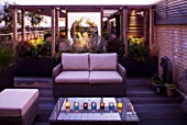 DESIGNER: CHARLOTTE ROWE  LONDON: ROOF GARDEN - A PLACE TO SIT - WICKER FURNITURE AND WOODEN PERGOLA WITH GLASS SCREEN LIT UP AT NIGHT,  DECKS, DECKING, SCREENING
