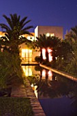 DESIGNERS ERIC OSSART AND ARNAUD MAURIERES  MOROCCO: AL HOSSOUN - COURTYARD SURROUNDING RECTANGULAR POOL AT NIGHT WITH LIGHTS FROM THE BUILDINGS