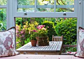 DESIGNER BUTTER WAKEFIELD  LONDON: VIEW OUT OF CONSERVATORY WITH CUSHIONS BY DRAGON FLOWER TO TERRACE/ PATIO WITH WOODEN TABLE AND CHAIRS