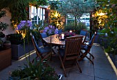 LONDON ROOFTOP GARDEN LIT UP AT NIGHT: WOODEN TABLE AND CHAIRS  OLEA EUROPEA  AGAPANTHUS  UMBELLATUS OVATA  LIMESTONE FLOOR