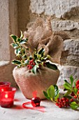 DESIGNER - JACKY HOBBS : CHRISTMAS DECORATION - CANDLES AND CHRISTMAS PUDDING WRAPPED IN HESIAN  HOLLY BERRIES AND LEAVES  IN WINDOWSILL
