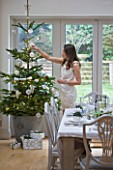 DESIGNER: JACKY HOBBS  LONDON - JACKY HOBBS HANGING BAUBLES ON CHRISTMAS TREE IN HER WHITE DINING ROOM