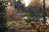 RHS GARDEN WISLEY  SURREY:  EVENING VIEW ACROSS THE LAKE AT SEVEN ACRES TO THE CHINESE PAGODA WITH HAMAMELIS APHRODITE  CAREX FLAGELLIFERA AND CAREX ASHIMENSIS EVERGOLD. WINTER