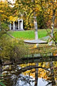 SALING HALL  ESSEX: AUTUMNAL VIEW ACROSS POND TO TEMPLE OF PISCES WITH HAMMOCK IN FIREGROUND SLUNG BETWEEN WHITE STEMS OF BIRCH - BETULA JERMYNS
