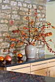 RICKYARD BARN HOUSE  OXFORDSHIRE: DESIGNERS JANE AND CLIVE NICHOLS. CHRISTMAS DECORATION ON WORKTOP IN KITCHEN - CONTAINER WITH MALUS BRANCHES  BAUBLES
