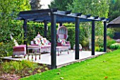 THE BOAT HOUSE: DESIGNER ARLETTE GARCIA - LAWN AND DECKED SEATING AREA WITH FURNITURE BY AMERICAN COMPANY MACKENZIE CHILDS - BLACK PERGOLA